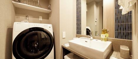 A compact hotel bathroom with a front-loading washing machine, a sink with a mirror above, and a towel rack.
