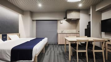 A hotel room with a bed, a desk, a chair, a television, a kitchen area, and a window.