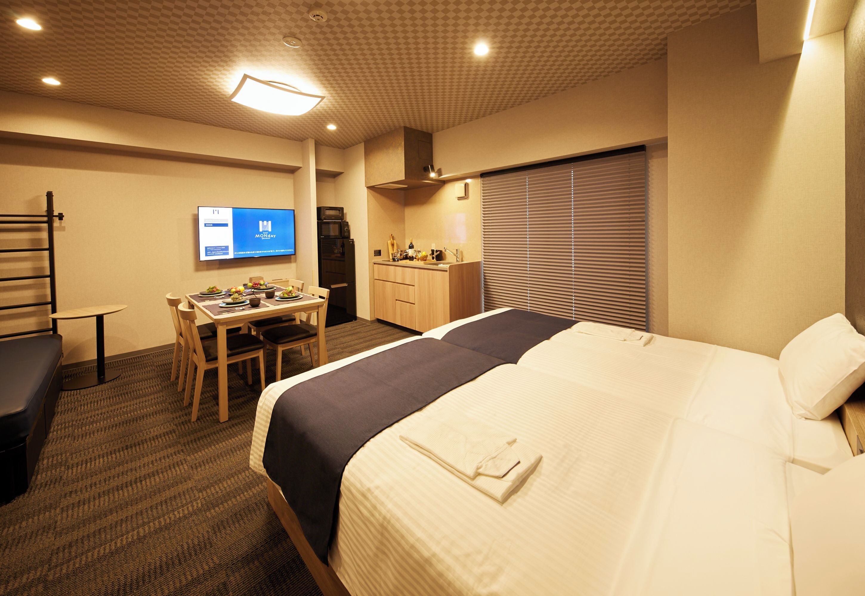A hotel room with a bed, a desk, a chair, a TV, and a kitchenette.