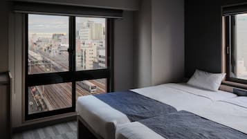 Une chambre d’hôtel dotée d’une grande fenêtre donnant sur la ville, d’un lit avec une literie blanche et grise, et d’une petite table de chevet.