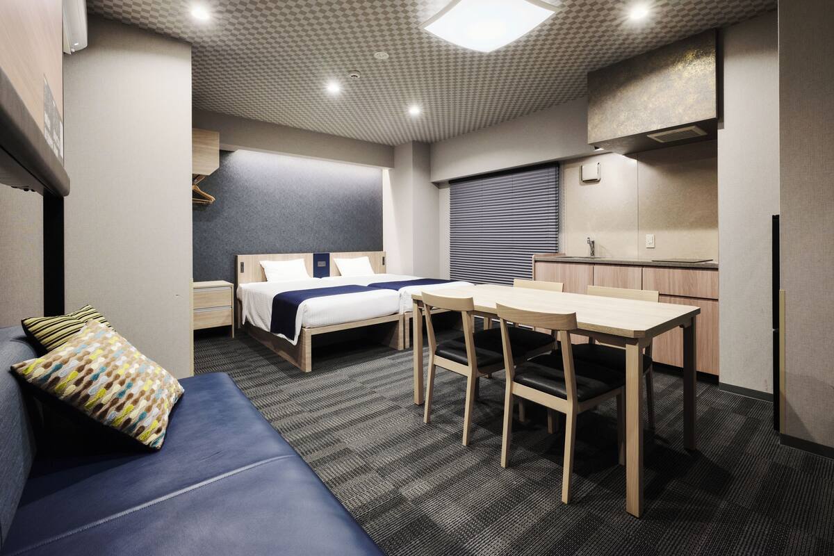 A compact hotel room with a bed, a desk, a chair, a sofa, and a kitchenette.