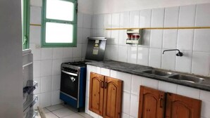 Une cuisine compacte avec des murs recouverts de carreaux blancs, une cuisinière à gaz, une machine à café et des placards en bois.