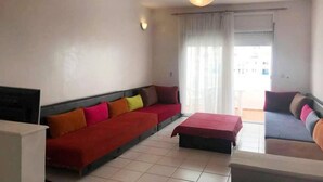 A living room with a red sofa, a blue sofa, and a red ottoman. There is a television on the left side and a window with curtains in the background.