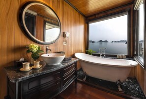 A bathroom with a freestanding bathtub, a round mirror, and a large window overlooking a body of water. - La Regina Legend Cruise (Hai Phong)