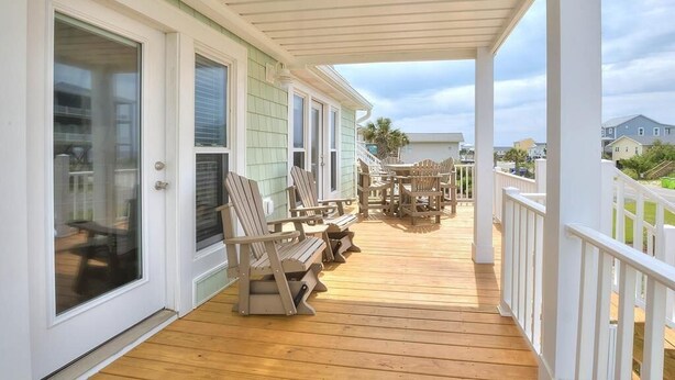 A covered porch with wooden flooring, white railings, and several Adirondack chairs.