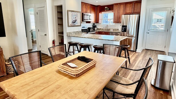 A modern kitchen with wooden cabinets, a central island, and a dining area with a wooden table and chairs. - Sun Catcher: Spacious 5BR Oak Island home with ocean views for 12 guests. (Oak Island)
