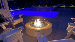 A poolside area with a circular fire pit surrounded by white lounge chairs.