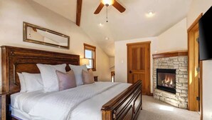 A bedroom with a large bed, a stone fireplace, wooden ceiling beams, and a ceiling fan.