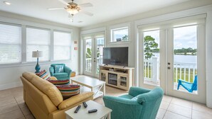 A living room with a beige sofa, teal armchairs, a coffee table, and a TV on a wooden stand. There are large windows and a door leading to a balcony with a view of a lake. - Fish, Beach, Pool, Swim, Relax in Luxury Rent for the month only 5,900.00 (Carrabelle)