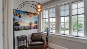 Een kamer met een groot raam, een schilderij van luchtballonnen, een stoel, een klein tafeltje en een vaas met bloemen.