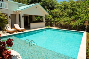 A rectangular swimming pool with clear blue water, surrounded by a white building with a light blue roof and a covered patio area with lounge chairs. - Large Oceanfront Villa with Pool, Close to Village (Placencia)