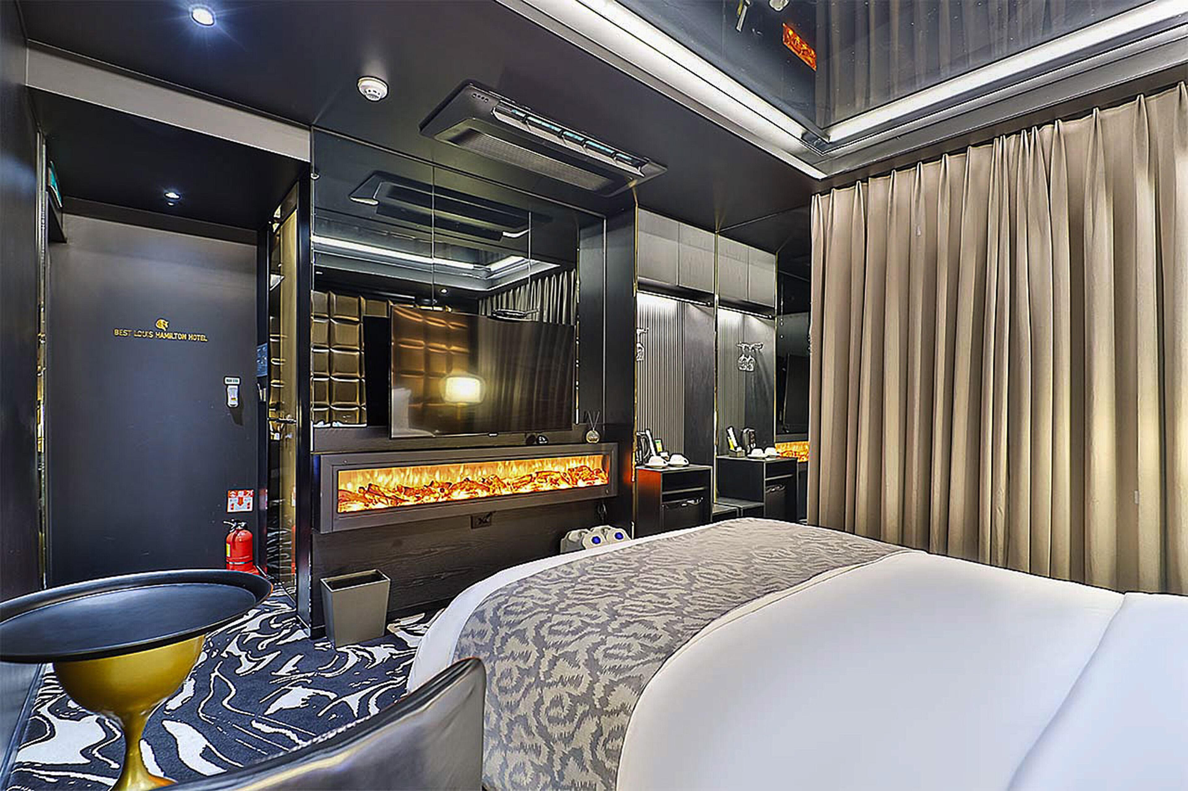 A modern hotel room with a large bed, a flat-screen TV, a fireplace, and a minibar.