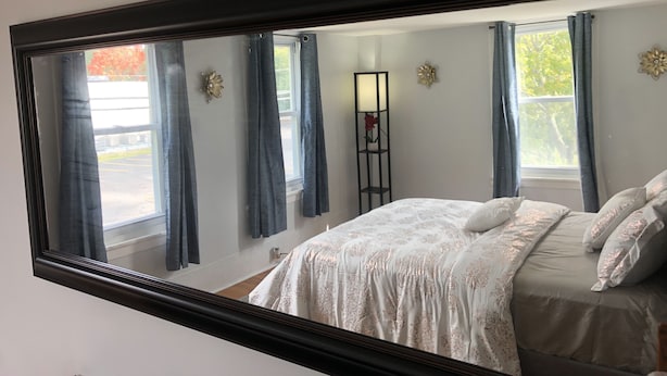 Un dormitorio con un espejo grande, una cama con ropa de cama floral, una lámpara de pie y una ventana con cortinas.