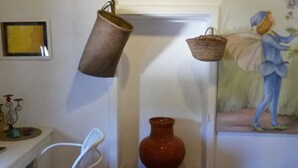 Ett rum med en vit vägg, en inramad tavla, en metallstol, en vit korg, en röd vas och en målning av en person.