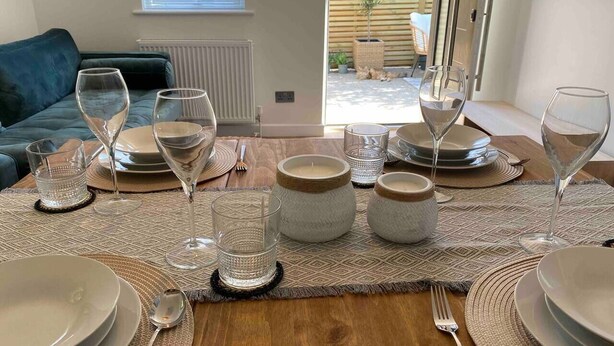 Une table à manger dressée avec des assiettes, des verres et des bougies, donnant sur un jardin grâce à une porte coulissante.