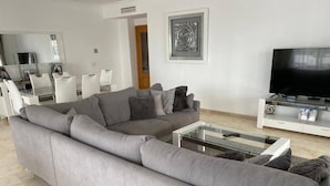 Een moderne woonkamer met een grijze hoekbank, een glazen salontafel, een flatscreen televisie, en een eethoek met witte stoelen en een glazen tafel.
