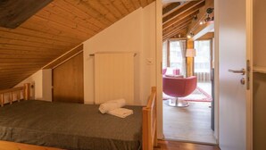 Une chambre avec un plafond en pente, un lit, une porte donnant sur une salle de bain, et une vue sur une chaise rose et une table dans la pièce adjacente.