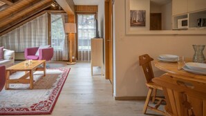 Une pièce chaleureuse de style loft, avec un plafond haut, des poutres en bois, un coin repas avec une table et des chaises, un canapé rose et une grande fenêtre avec des rideaux.