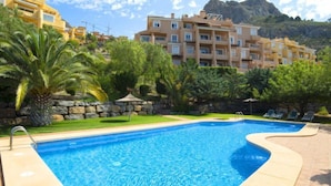 A swimming pool with a curved edge, surrounded by a stone deck. In the background, there are multi-story buildings with balconies and a mountainous landscape. - Residencial Jazmines by Interhome (Calpe/Calp)