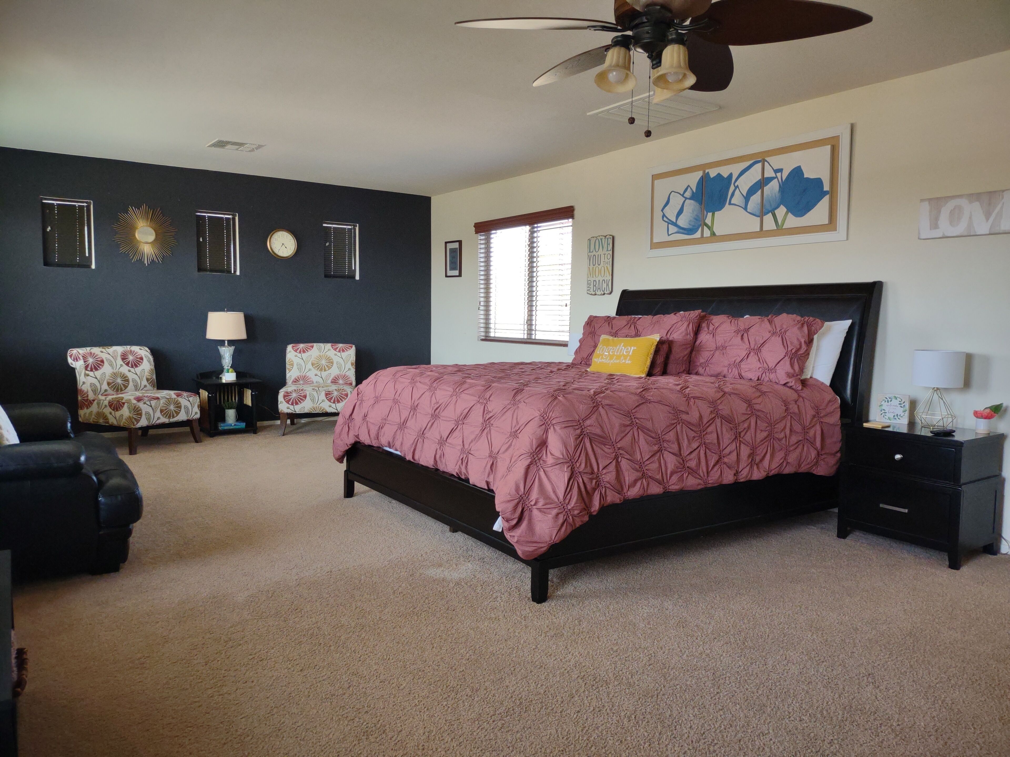 A bedroom with a bed, two chairs, a nightstand, a ceiling fan, and wall decorations.