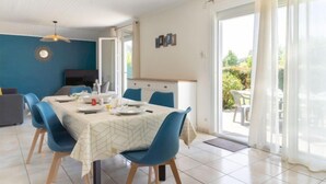 A dining area with a table set for four, blue chairs, a ceiling light fixture, and a view of the outdoors through a glass door. - Les Trounques by Interhome (Mimizan)