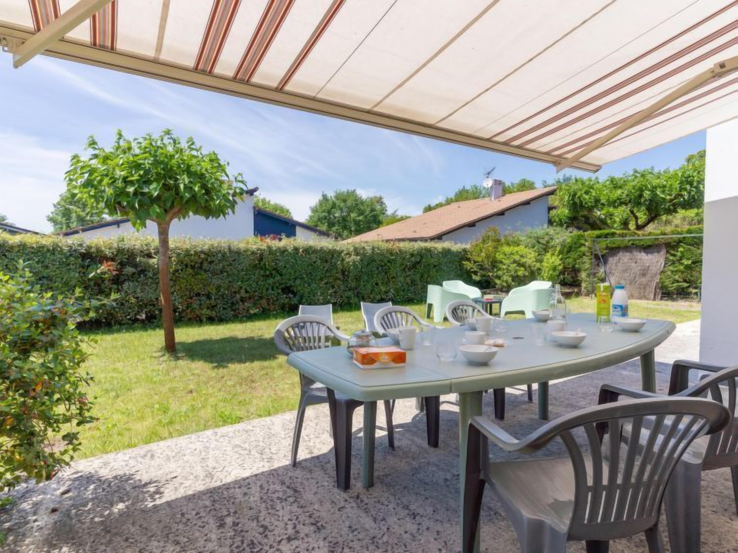 An outdoor patio area with a table set for four, surrounded by chairs, under a shaded canopy.