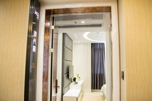 Een moderne hotelkamer met een houten deur, een flatscreen televisie, een klein tafeltje met een vaas en een groot raam met gordijnen.