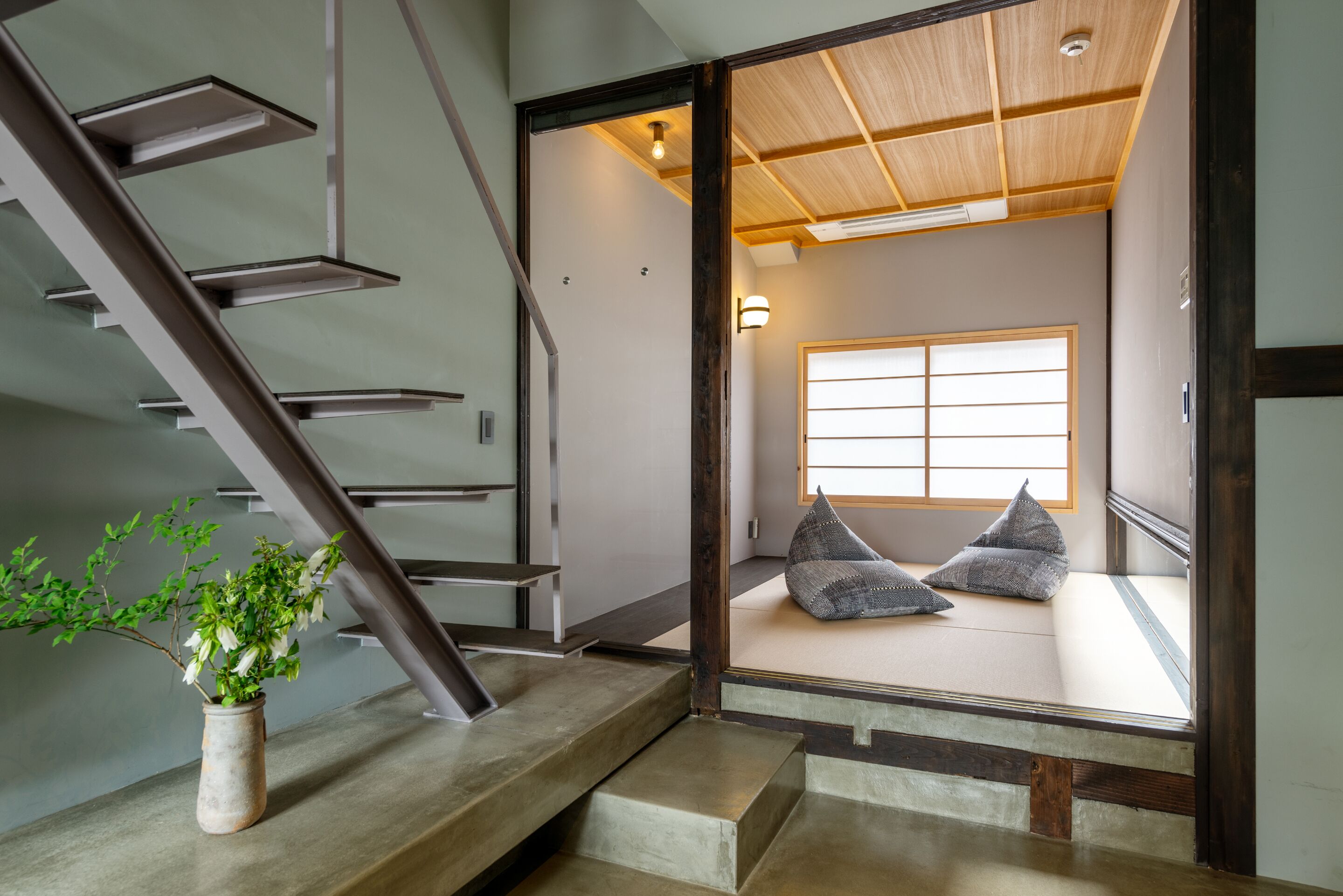 A traditional Japanese interior with tatami flooring, a wooden staircase, and a view of a room with tatami mats and cushions.
