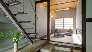 Un intérieur japonais traditionnel avec un sol recouvert de tatamis, un escalier en bois et une vue sur une pièce également dotée de tatamis et de coussins.