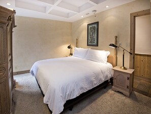 A bedroom with a large bed, two bedside tables, a nightstand, a framed picture on the wall, and a wooden wardrobe. - Village Center Condo 1F (Vail)