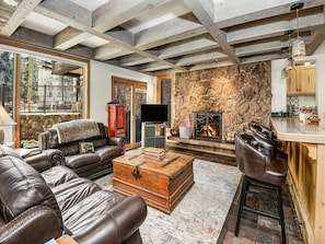 A cozy living room with a stone fireplace, leather furniture, and a rustic kitchen area. - Village Center Condo 1F (Vail)