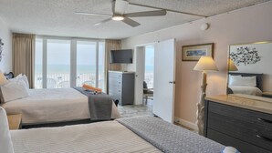 Habitación de hotel con dos camas, ventilador de techo, televisor, escritorio con lámpara y vista al océano.