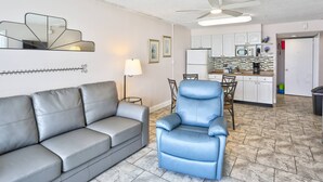 Un espacio habitable compacto con un sofá gris, un sillón azul, una zona de comedor con mesa y sillas, una kitchenette con microondas y un ventilador de techo.
