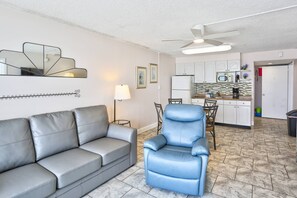 A compact living space with a grey sofa, a blue armchair, a dining area with a table and chairs, a kitchenette with a microwave, and a ceiling fan.