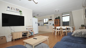 A modern living room with a flat-screen TV, wooden furniture, a dining area with a table and chairs, and a kitchen in the background. - Relaxed, beachy 2 bed apartment, only 1.9kms from the beach, close to shops (Kingscote)