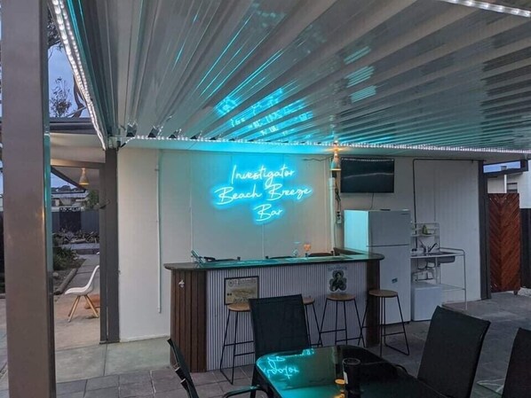 An outdoor bar area with a neon sign reading "Investigator Beach Breeze Bar," a bar counter, and seating with neon-lit chairs.