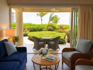 Una habitación con sofá, dos sillones, una mesa con un florero y vistas a un jardín cuidado con palmeras.