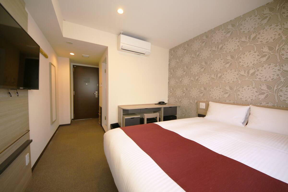 A hotel room with a bed, a desk, a television, and an air conditioning unit.