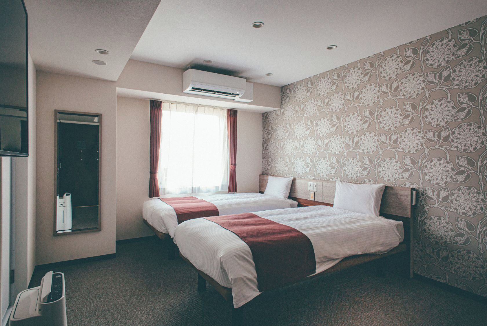 Habitación de hotel con dos camas, aire acondicionado, un espejo y un papel tapiz estampado.