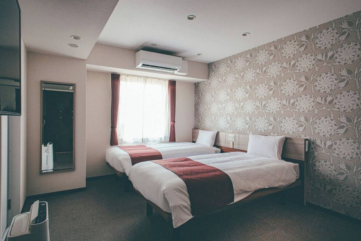 A hotel room with two beds, an air conditioning unit, a mirror, and a patterned wallpaper.