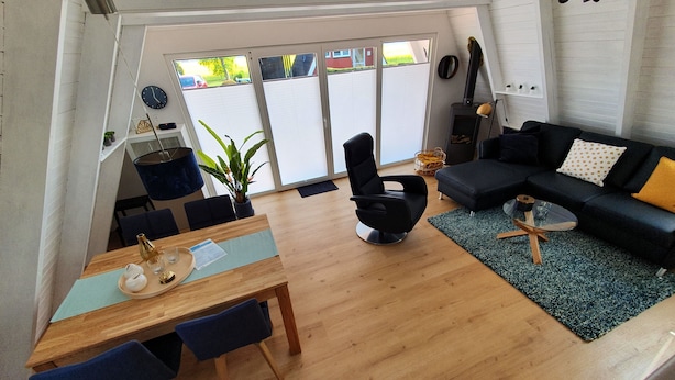 Ein modernes Wohnzimmer mit einer schwarzen Couch, einem Esstisch mit Stühlen, einem Couchtisch und einem großen Fenster mit Jalousien.