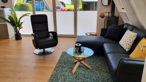 Ein modernes Wohnzimmer mit einer schwarzen Ledersofa, einem schwarzen Sessel und einem Glastisch. Eine Topfpflanze und ein kleiner Tisch mit einer Lampe stehen dort. Große Fenster bieten einen Ausblick nach draußen.