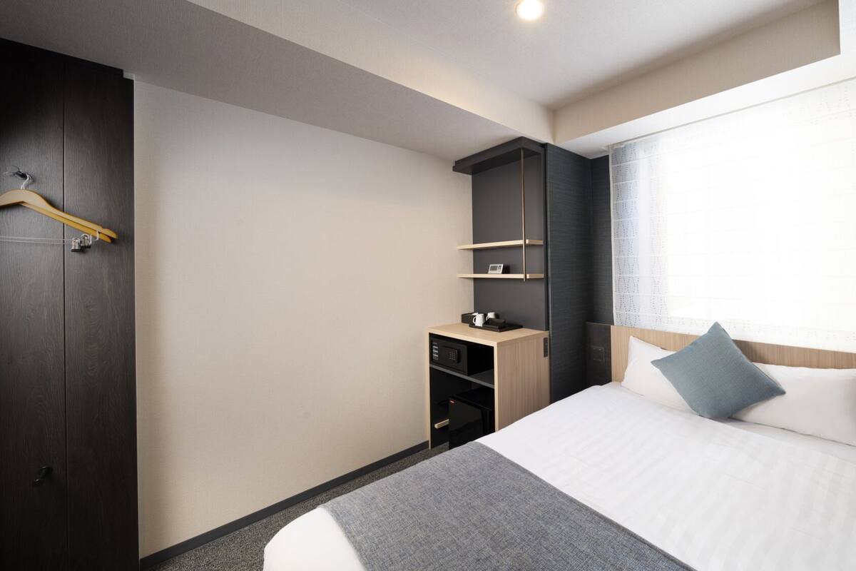 Une chambre d’hôtel avec un lit, une armoire, un bureau avec un téléphone et une fenêtre avec des rideaux.