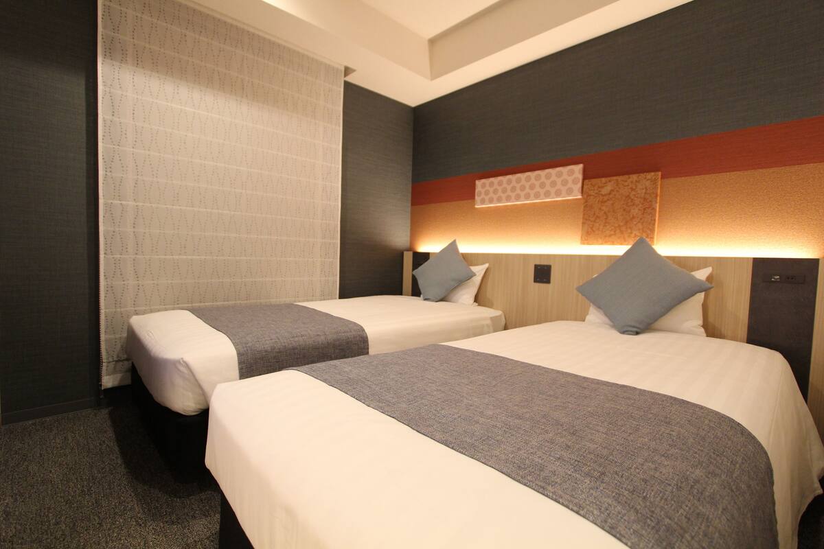 Une chambre d’hôtel avec deux lits, une grande fenêtre avec des rideaux à motifs et une tête de lit moderne dotée d’un éclairage intégré.