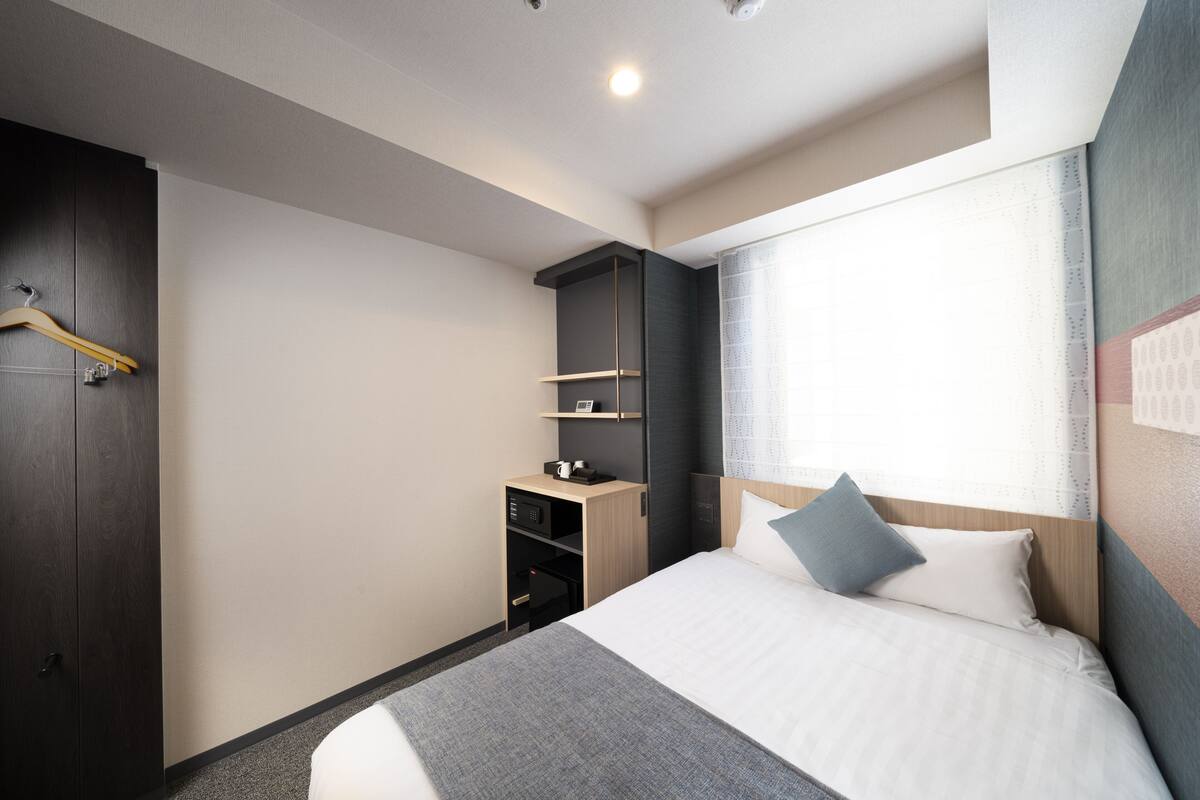 Une chambre d’hôtel moderne avec un lit bien fait, un petit bureau avec une chaise, une armoire et une fenêtre avec des rideaux.