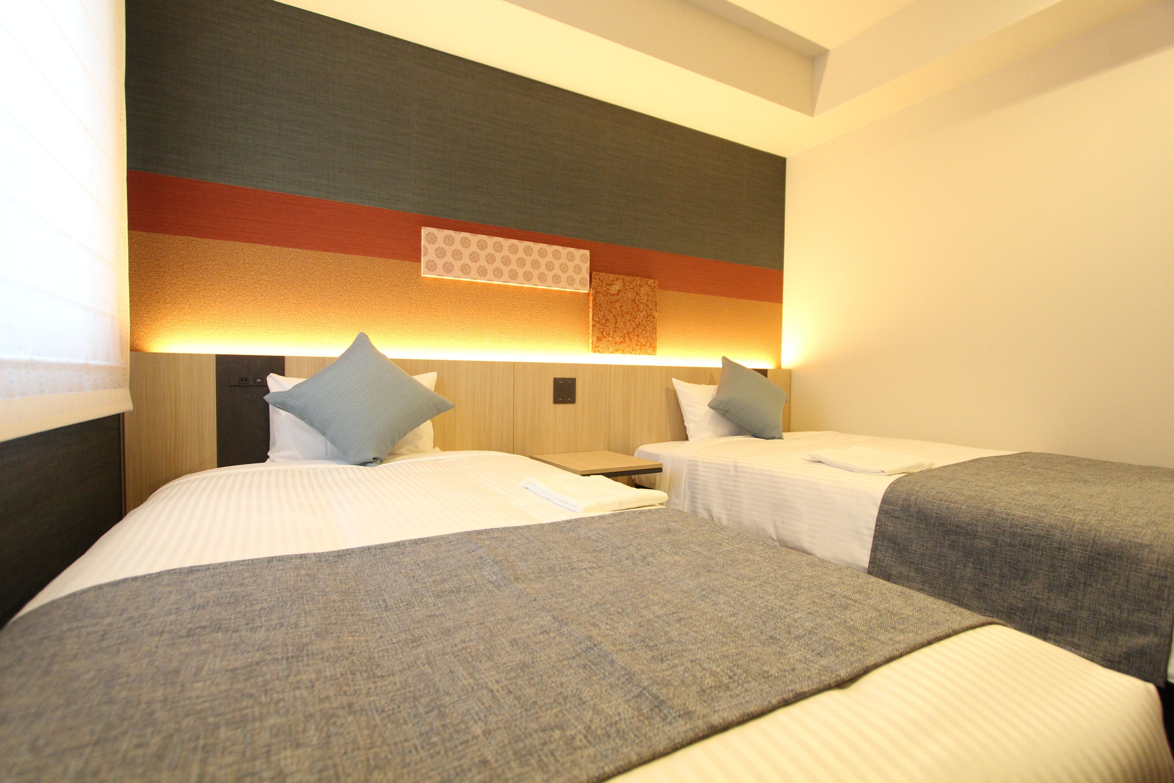 A hotel room with two beds, each with a pillow and a blanket. There is a nightstand between the beds, and a wall-mounted lamp above the beds.