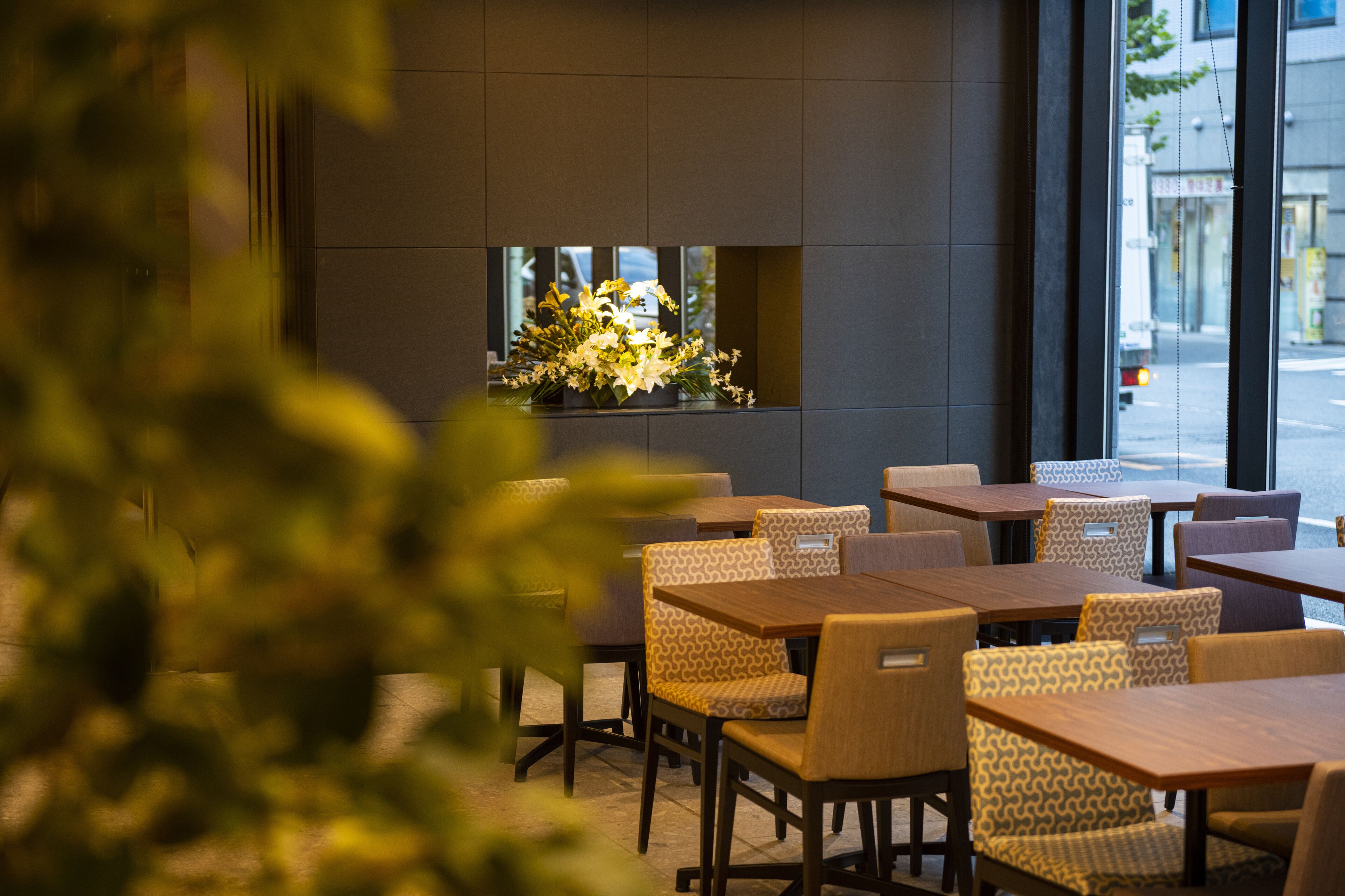 L'intérieur d'un café moderne, avec des tables en bois et des chaises à motifs, une fenêtre agrémentée d'un arrangement floral et une vue sur la rue.