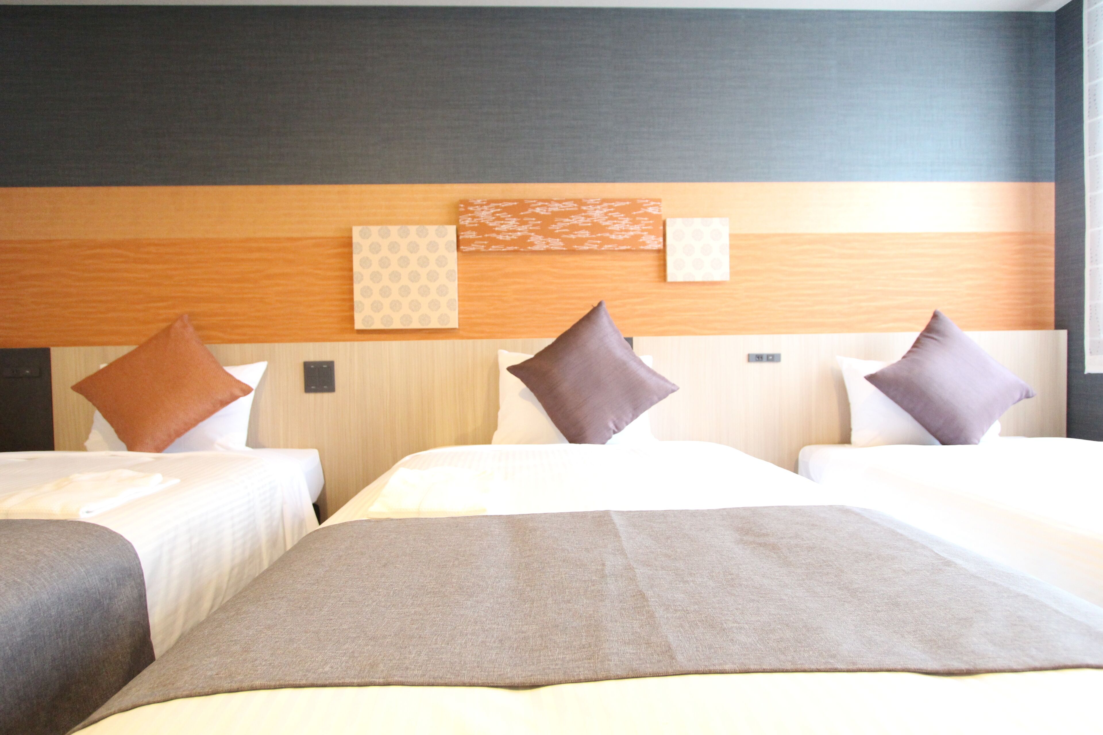 A hotel room with two beds, each with a brown blanket and multiple pillows. The headboard has a wooden panel with a geometric pattern.
