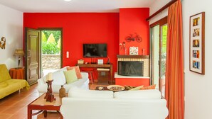 A living room with a red wall, a fireplace, a television, and a dining area with a table and chairs. - Villas Arhontika Red, Nestled in Chestnut Forest in Cretan Mountainside (Elos)