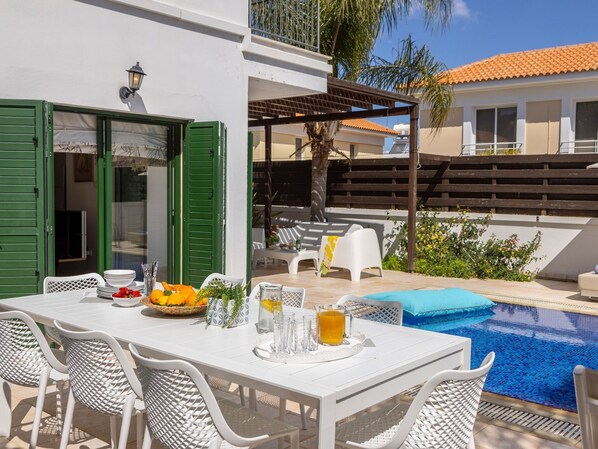 Un área de comedor junto a la piscina con una mesa lista para dos, una piscina pequeña y un espacio de descanso cubierto.